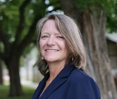 Headshot of and older white woman with shoulder length hair wearing a dark blue shirt. She's outside standing in front of a tree.